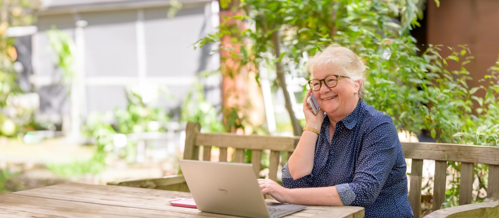 Smiling woman talking on the phone, using a laptop outdoors.