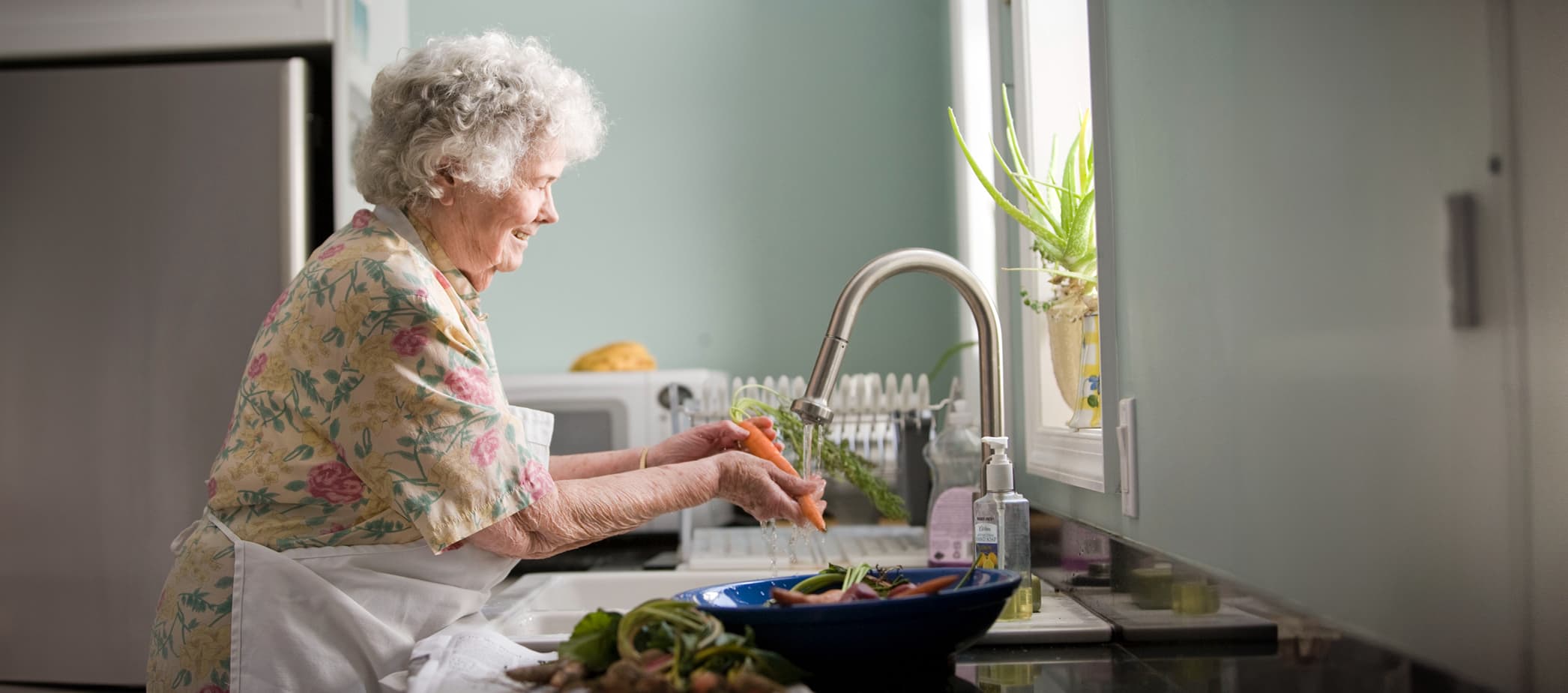 Elderly woman washing vegetables in a bright kitchen.