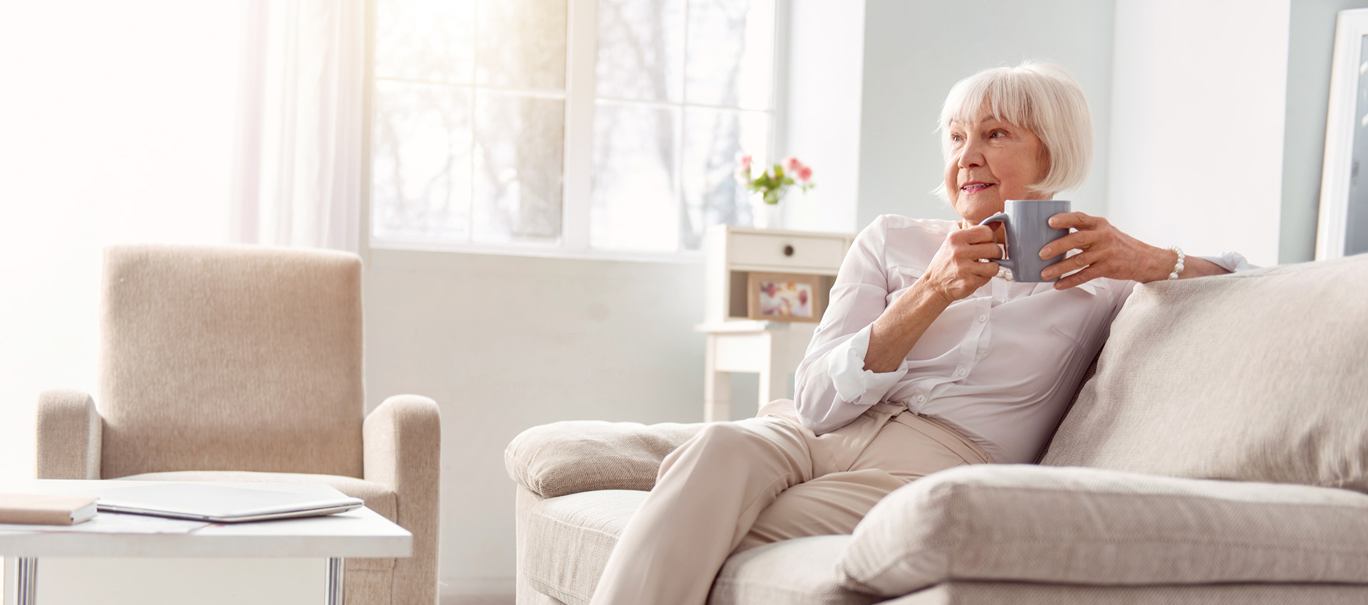 senior woman sitting on the couch in her living room, drinking coffee and contemplating something